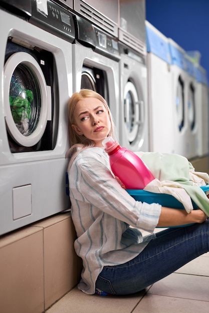 Ama de casa cansada de 20 años que lleva la cesta de la ropa sucia con ropa sucia esperando el final del lavado, sentada en el suelo con mal humor, agotada. en la casa de lavado