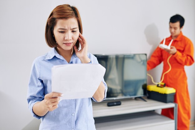 Ama de casa con el ceño fruncido mirando la factura del servicio del técnico que instala el enrutador wi-fi en su apartamento