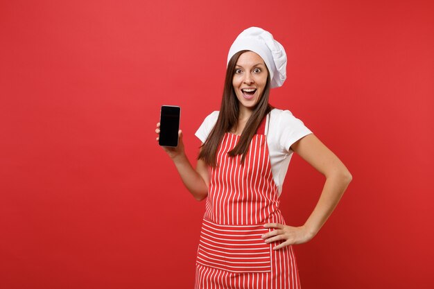 Ama de casa cocinera cocinera o panadero en delantal de rayas camiseta blanca gorro de chef de toque aislado sobre fondo de pared roja. Mujer mantenga la pantalla en blanco del teléfono móvil para contenido promocional Mock up copy space concept