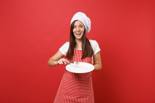 Ama de casa cocinera cocinera o panadero en delantal de rayas, camiseta blanca, sombrero de chef toque aislado sobre fondo de pared roja. Mujer ama de llaves mantenga plato redondo vacío con un tenedor. Burlarse del concepto de espacio de copia.