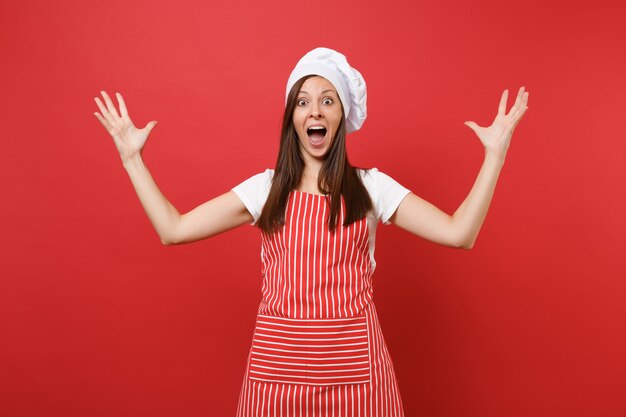 Ama de casa cocinera cocinera o panadero en delantal de rayas, camiseta blanca, sombrero de chef toque aislado sobre fondo de pared roja. Mujer feliz ama de llaves expresiva extendiendo las manos. Burlarse del concepto de espacio de copia.