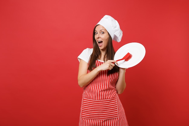 Ama de casa cocinera cocinera o panadero en delantal de rayas, camiseta blanca, sombrero de chef toque aislado sobre fondo de pared roja. Mujer mantenga plato blanco con cepillo para lavar platos. Burlarse del concepto de espacio de copia.