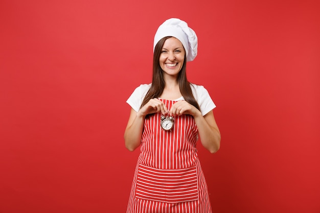 Ama de casa cocinera cocinera o panadero en delantal de rayas, camiseta blanca, sombrero de chef toque aislado sobre fondo de pared roja. Mujer sonriente sostenga en la mano el despertador retro date prisa. Burlarse del concepto de espacio de copia.