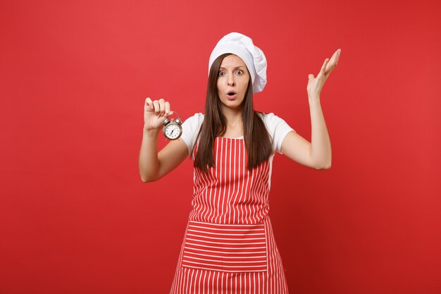 Ama de casa cocinera cocinera o panadero en delantal de rayas, camiseta blanca, sombrero de chef toque aislado sobre fondo de pared roja. Mujer sorprendida sostenga en la mano el despertador retro date prisa. Burlarse del concepto de espacio de copia.