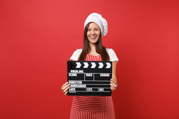 Ama de casa cocinera cocinera o panadero en delantal de rayas, camiseta blanca, sombrero de chef toque aislado sobre fondo de pared roja. Mujer sosteniendo claqueta de cine negro clásico. Burlarse del concepto de espacio de copia.