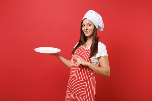 Ama de casa cocinera cocinera o panadero en delantal de rayas, camiseta blanca, sombrero de chef toque aislado sobre fondo de pared roja. La mujer sostiene un plato redondo en blanco vacío con lugar para la comida. Simulacros de concepto de espacio de copia