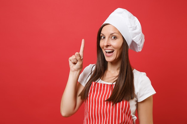 Ama de casa cocinera cocinera o panadero en delantal de rayas rojas, camiseta blanca, sombrero de chef de toque aislado sobre fondo de pared roja. Retrato de mujer morena ama de llaves de cerca. Burlarse del concepto de espacio de copia.