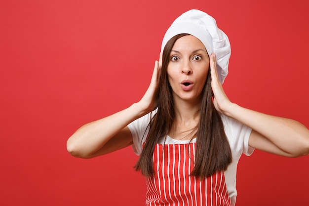 Ama de casa cocinera cocinera o panadero en delantal de rayas rojas, camiseta blanca, sombrero de chef de toque aislado sobre fondo de pared roja. Retrato de mujer morena ama de llaves de cerca. Burlarse del concepto de espacio de copia.