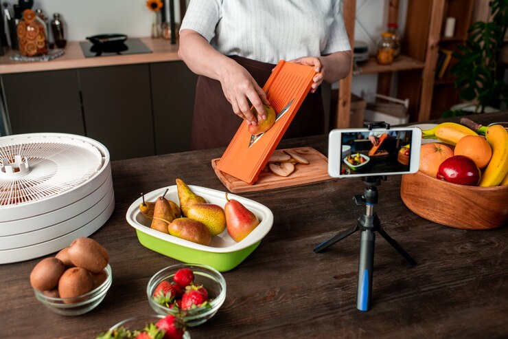 Foto ama de casa contemporánea o bloguera de pie junto a la mesa de la cocina frente a la cámara del teléfono inteligente y grabando video de hacer frutas secas