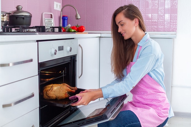 Ama de casa en delantal horneando pollo en el horno para cenar en la cocina
