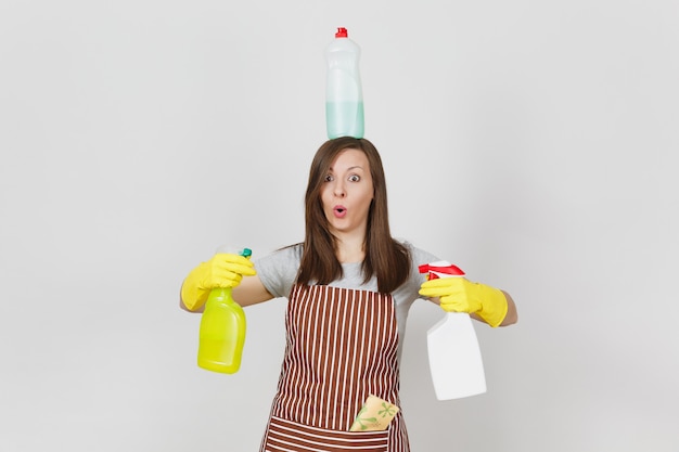 Ama de casa divertida en guantes amarillos, delantal de rayas, trapo de limpieza en el bolsillo sobre fondo blanco. Mujer sosteniendo en las manos, en la cabeza botellas con líquido limpiador para lavar los platos. Copie el espacio para publicidad.