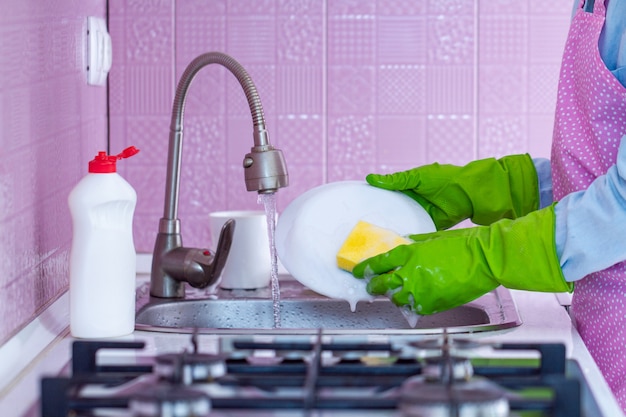 Ama de casa con guantes de goma y delantal lava platos en casa con esponja y detergente