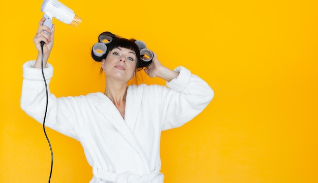 Ama de casa hermosa joven en una túnica blanca. rulos en la cabeza. Fondo amarillo. seque su cabello con un secador de pelo. Baner, lugar para el texto