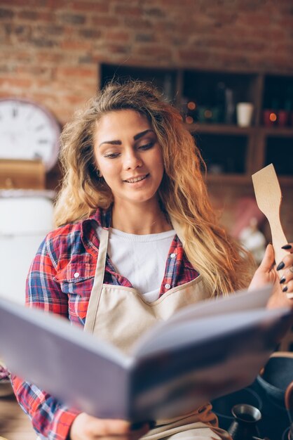 Ama de casa joven en un delantal lee el libro de recetas