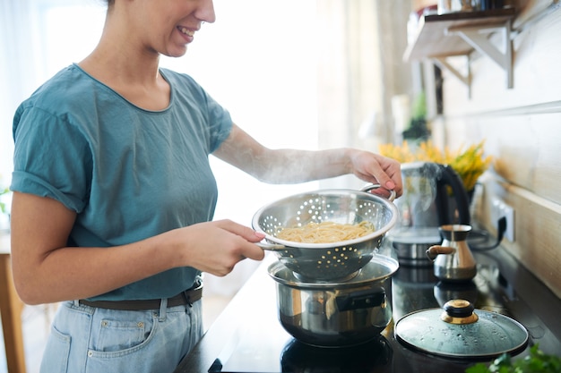 Ama de casa joven con espaguetis cocidos calientes en un colador de pie junto a la estufa eléctrica en la cocina