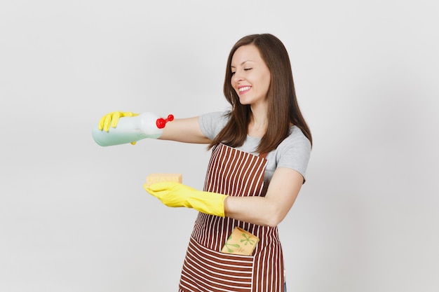 Ama de casa joven en guantes amarillos, delantal rayado, trapo de limpieza en el bolsillo aislado sobre fondo blanco. La mujer vierte un líquido más limpio para lavar los platos de la botella en una esponja. Copie el espacio para publicidad.