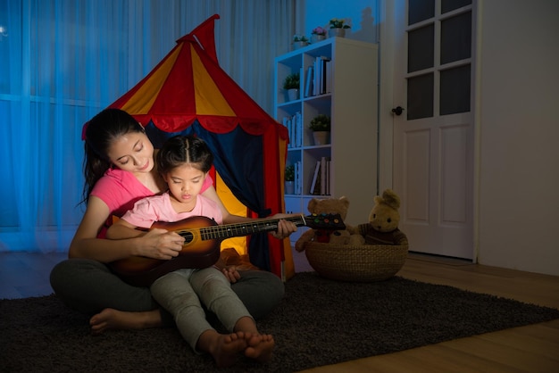 Ama de casa joven y tranquila enseñando a una hermosa hija tranquila tocando una pequeña guitarra de ukelele sentada en el suelo de la sala de estar junto con una carpa de niños de juguete por la noche.