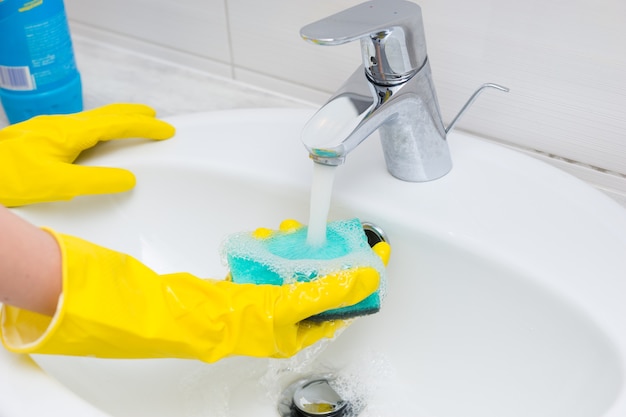 Ama de casa lavando el lavabo en el baño con sus manos enguantadas enjuagando una esponja bajo el agua que fluye del grifo o grifo, cerca de sus manos