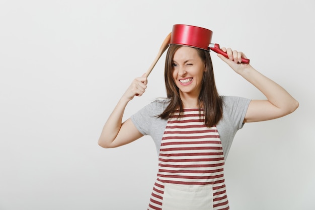 Ama de casa loca sonriente joven en delantal rayado, camiseta gris aislada. Hermosa mujer de ama de llaves divertida sosteniendo una cacerola vacía roja en la cabeza cuchara de madera