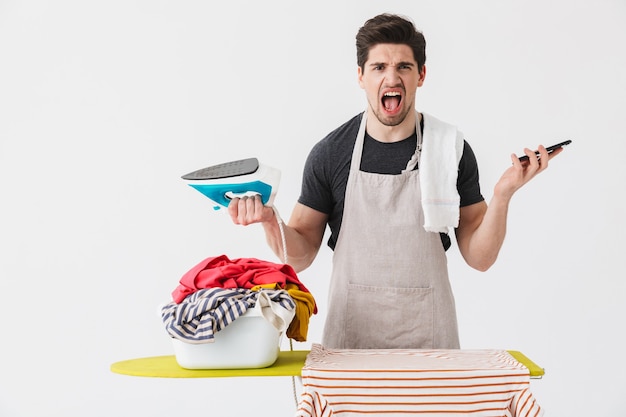 Ama de casa morena enojado guapo vistiendo delantal que se encuentran aisladas sobre blanco, planchando ropa en una tabla, hablando por teléfono móvil