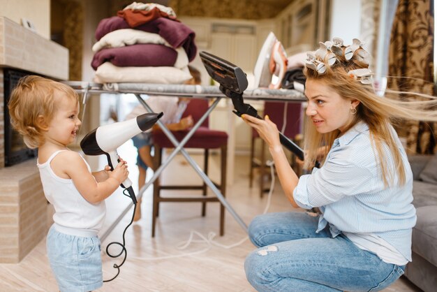 Ama de casa con niño jugando aspiradora y secador de pelo en la tabla de planchar. Mujer con niño haciendo quehaceres domésticos en casa juntos. Persona de sexo femenino con hija divirtiéndose en su casa