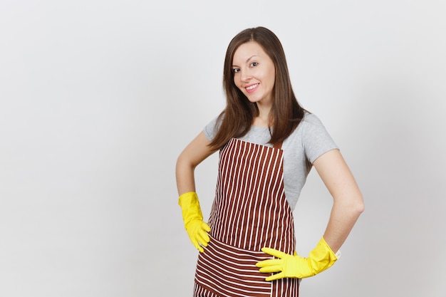 Ama de casa sonriente atractiva joven en delantal rayado, guantes amarillos aislados. Mujer hermosa ama de llaves de pie con los brazos en jarras