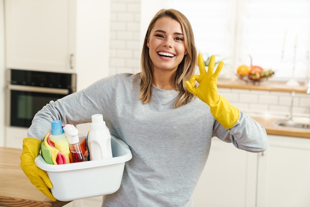 Ama de casa sonriente joven en guantes sosteniendo botellas de limpiador y mostrando el signo de ok en la cocina moderna