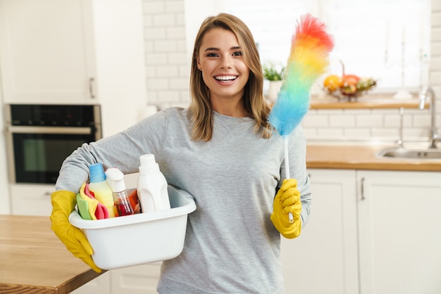 Ama de casa sonriente joven en guantes sosteniendo botellas de limpiador y plumero de colores en la cocina moderna