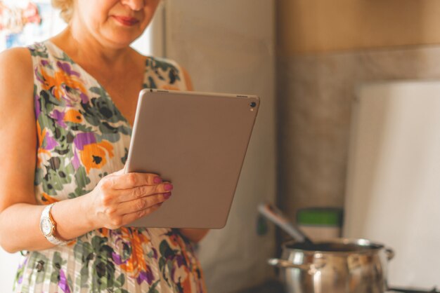 Ama de casa sosteniendo un teléfono inteligente o tableta para verificar el recibo mientras cocina en la cocina