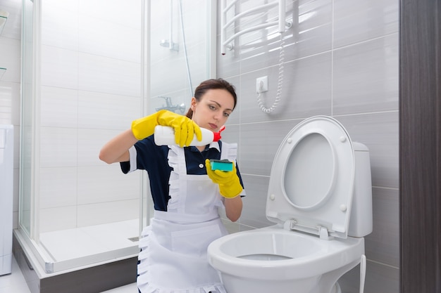 Ama de llaves femenina en vestido azul y delantal blanco poniendo limpiador en una esponja verde en preparación para fregar el inodoro en el baño