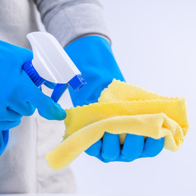 El ama de llaves de la mujer joven está limpiando la mesa blanca en el delantal con los guantes azules, limpiador en aerosol, trapo amarillo mojado, cierre para arriba, espacio de la copia, concepto de diseño en blanco.