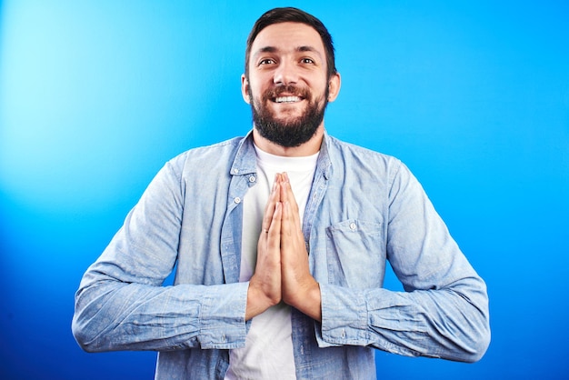 Foto amable amable hombre barbudo caucásico sonriendo con las manos cruzadas en oración agradecida pose aislado sobre fondo azul.