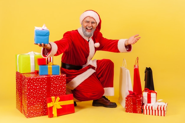 Amable anciano con barba gris vistiendo traje de santa claus sentado rodeado de cajas de regalo y bolsas de compras, saludo con año nuevo. Tiro de estudio interior aislado sobre fondo amarillo.