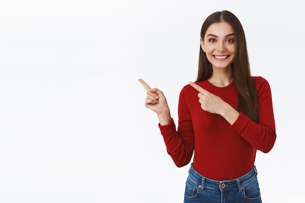 Amable, asertiva chica guapa morena mostrando con gusto la promoción del cliente, buena oferta, señalando la esquina superior izquierda, sonriendo feliz, ayudando a la persona que pide dirección, de pie fondo blanco