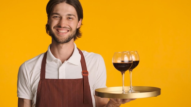 Amable camarero sosteniendo una bandeja con copas de vino sobre un fondo colorido Joven barbudo en delantal sonriendo a la cámara