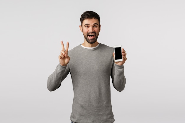 Amable y despreocupado, divertido, hombre gay con barba en suéter gris, edite la foto de perfil de la aplicación de citas en línea, haga el signo de la paz, muestre la pantalla del teléfono inteligente y sonría feliz