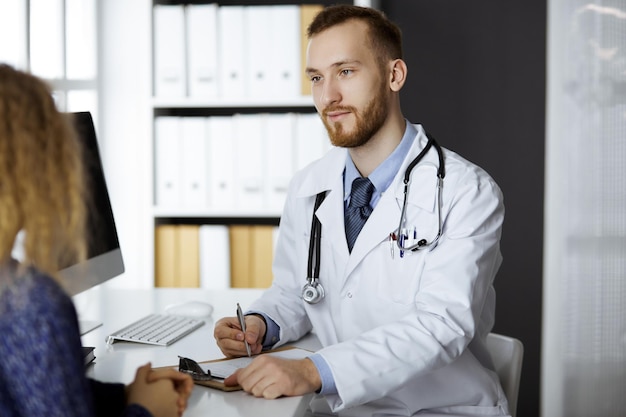 Amable doctor de barba roja y mujer paciente discutiendo el examen de salud actual mientras está sentado en la clínica. Servicio médico perfecto y concepto de medicina.