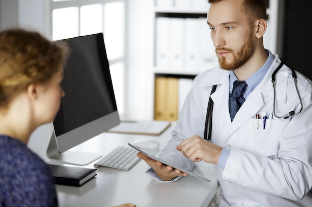 Amable doctor de barba roja y mujer paciente discutiendo el examen de salud actual mientras se sienta y usa una tableta en la clínica. concepto de medicina.