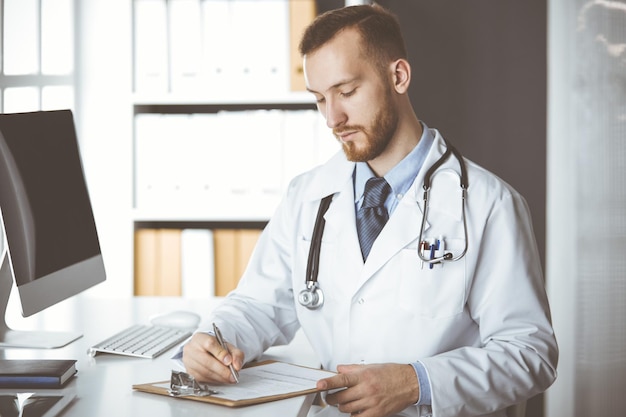 Amable doctor de barba roja sentado y escribiendo en el portapapeles en la clínica. Perfecto servicio médico en el hospital. concepto de medicina.