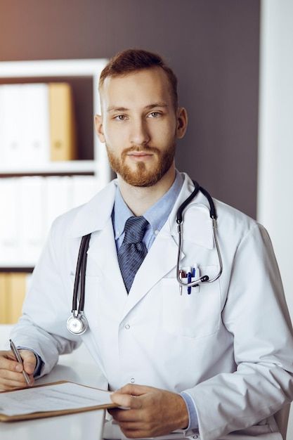 Amable doctor de barba roja sentado y escribiendo en el portapapeles en una clínica soleada. concepto de medicina.