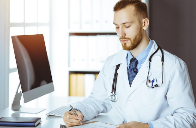 Amable doctor de barba roja sentado y escribiendo en el portapapeles en una clínica soleada. concepto de medicina.