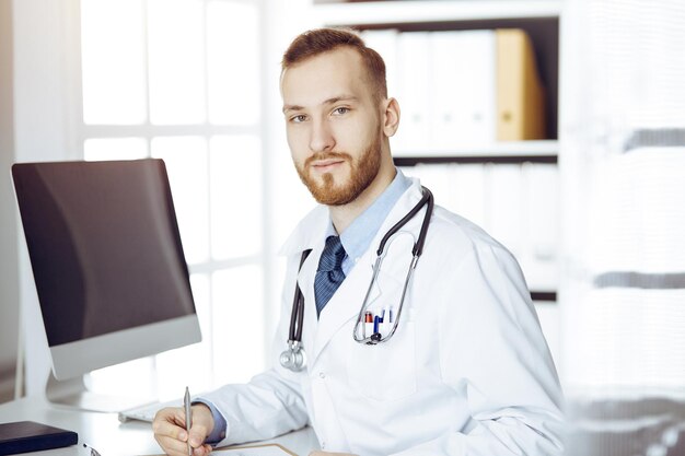 Amable doctor de barba roja sentado y escribiendo en el portapapeles en una clínica soleada. concepto de medicina.