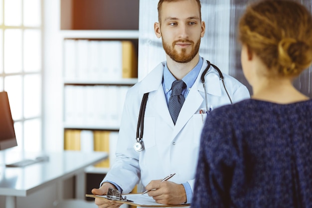 Amable doctor barbudo y mujer paciente discutiendo el examen de salud actual mientras está sentado en la soleada clínica Concepto de medicina