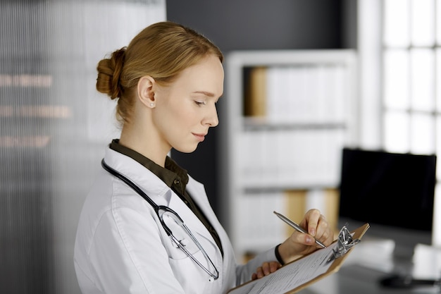 Amable doctora sonriente usando portapapeles en la clínica. Retrato de una mujer médica amigable en el trabajo. Servicio médico en el hospital. Concepto de medicina.