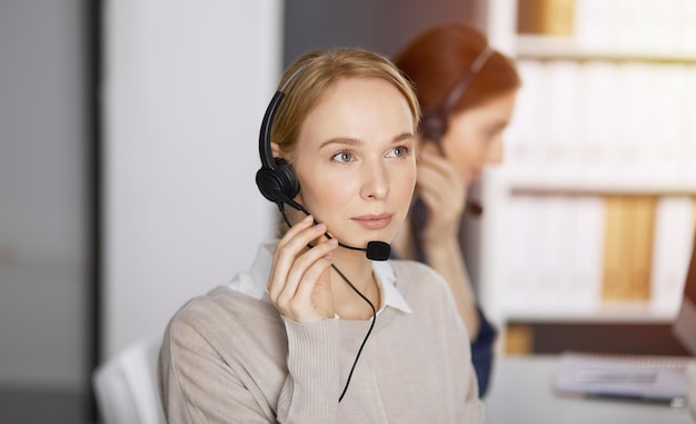 Amable empresaria caucásica hablando por auriculares en la oficina soleada. Centro de llamadas y grupo de personas diversas en los negocios.