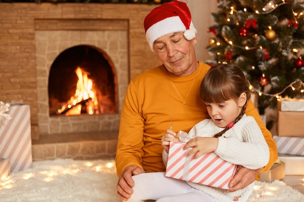 Amable hombre mayor vestido con suéter naranja y sombrero de santa claus posando con niña abriendo su regalo de Navidad con expresión facial sorprendida. Feliz navidad y próspero año nuevo.