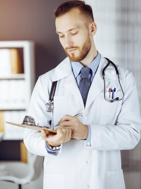 Amable médico de barba roja de pie y escribiendo con portapapeles en una clínica soleada en su lugar de trabajo. Concepto de medicina.