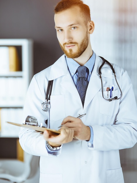 Amable médico de barba roja de pie y escribiendo con portapapeles en una clínica soleada en su lugar de trabajo. Concepto de medicina.