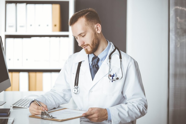 Amable médico de barba roja de pie y escribiendo con portapapeles en la clínica en su lugar de trabajo. Perfecto servicio médico en el hospital. Concepto de medicina.