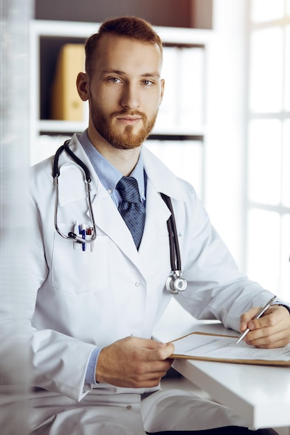 Amable médico barbudo sentado y escribiendo en el portapapeles en una clínica soleada. Concepto de medicina.